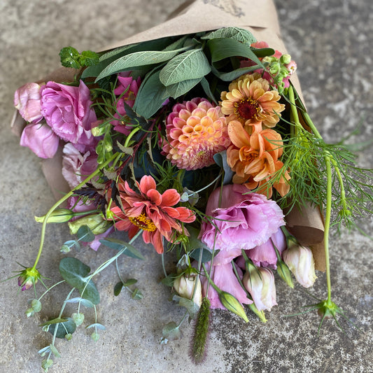A freshly cut summer bouquet featuring a variety of flowers including pink roses, snapdragons, and other assorted plants, wrapped in a brown paper bag.